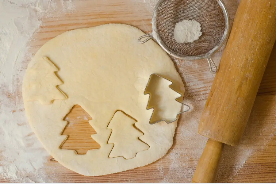 “Close-up of a baking scene with cookie dough rolled out, tree-shaped cookie cutters, a wooden rolling pin, and a dusting of flour on a wooden surface.”