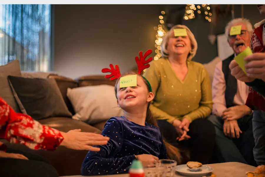 “A family gathered in a cosy living room, laughing while playing a guessing game with sticky notes on their foreheads. A young girl with festive reindeer antlers smiles as she participates.”