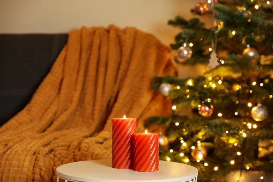 “A warm, inviting corner of a living room with two lit red candles on a table, a soft throw blanket draped over a chair, and a decorated Christmas tree glowing in the background.”