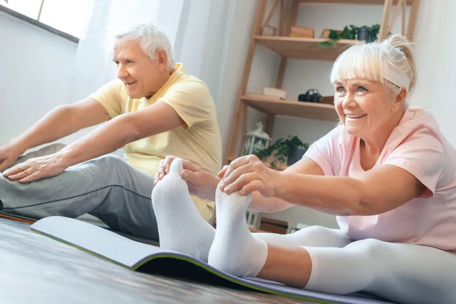 An elderly couple, male and female, sit on the floor on grey exercise mats, stretching to try and reach their toes.