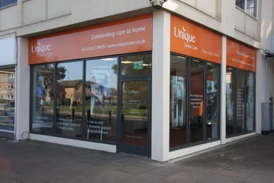 The front of Unique Senior Care shop unit. It is a corner unit with an orange banner at the top, with white writing which reads: 'Unique Senior Care. Outstanding care at home.'