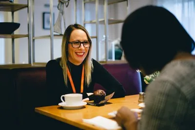 Two women sit at a table with cups of tea, talking. One of them women has an orange Unique Senior Care lanyard on. The other has her back to the camera.