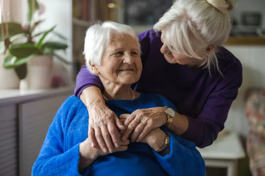 An older woman in a blue sweater smiles as a younger woman in purple leans over and hugs her in a cosy home setting.