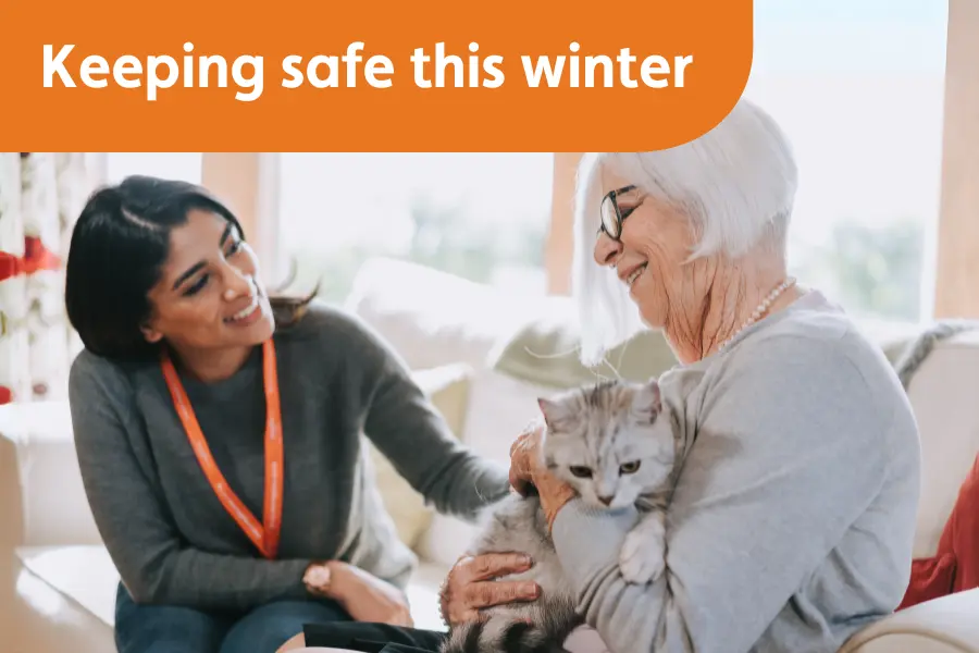 A lady and caregiver sat together smiling holding a cat, overlayed with the text 'keeping safe this winter'