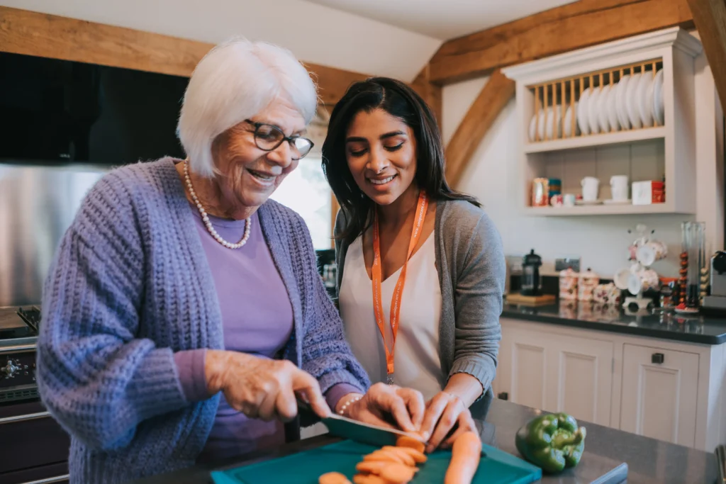 An older lady and her caregiver smiling while cooking a meal together in the kitchen