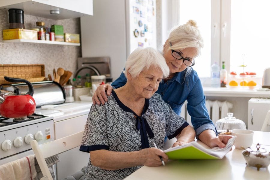 A senior woman sits at a kitchen counter with a lady dressed in blue standing beside her. They are both reading from a green folder potentially on the subject of live in care costs