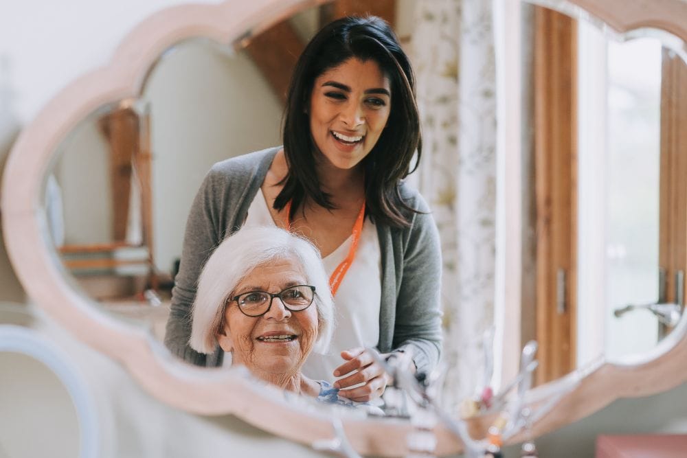 Two ladies stand in front of a mirror. An elderly lady sits smiling, a younger lady stands behind her smiling. The younger lady wears an orange lanyard.