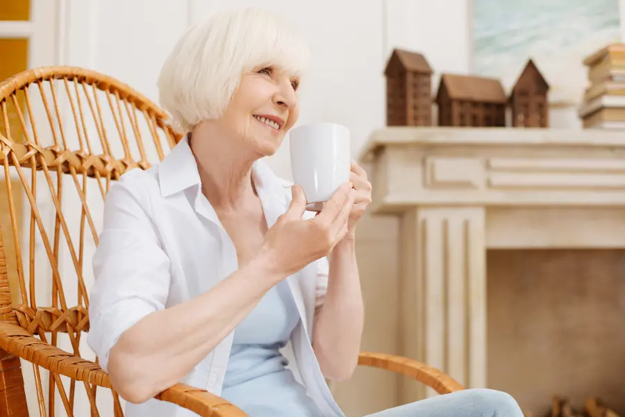 Smiling elderly woman with short blonde hair sitting in a wicker chair, holding a white mug.