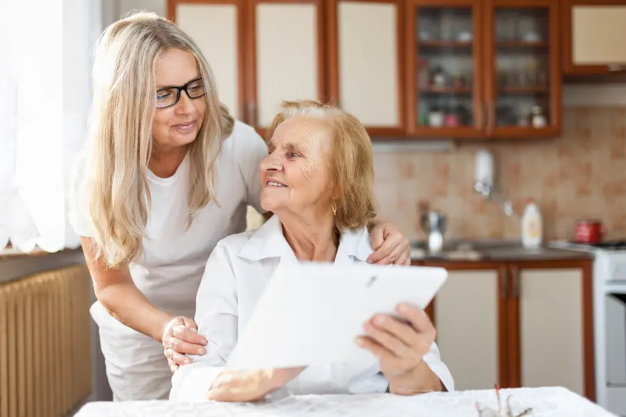 A lady stood behind an older woman sitting at a table, both looking at something on an Ipad