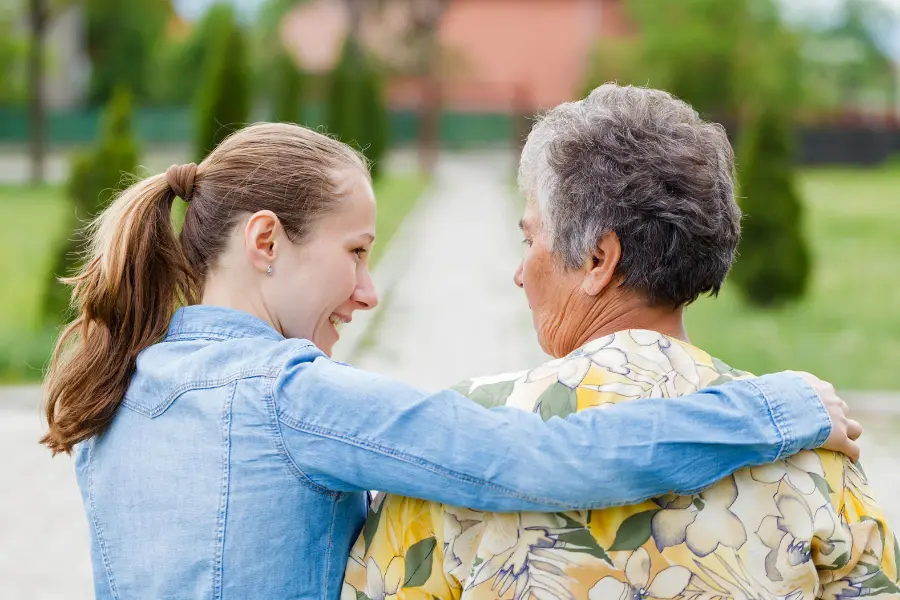 Young woman with her arm around an elderly woman as they walk outdoors, seen from behind.