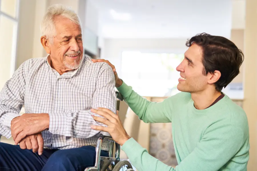 Older man in a wheelchair smiling at a younger man, who is kneeling and has a hand on the older man's shoulder.