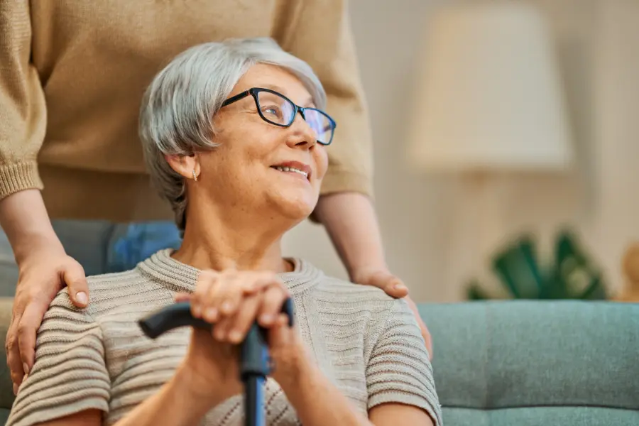 Elderly woman with grey hair and glasses, smiling and holding a cane, with a person standing behind her with hands on her shoulders.