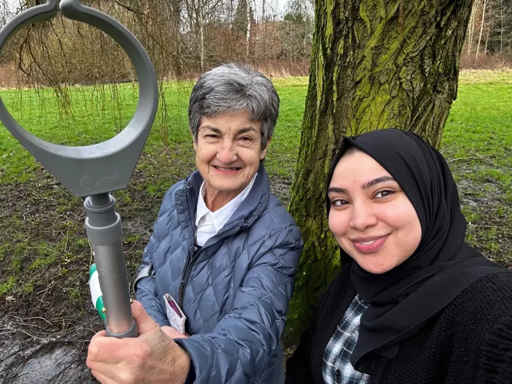 Older woman with grey hair and a younger woman in a headscarf, both smiling and taking a selfie outdoors.