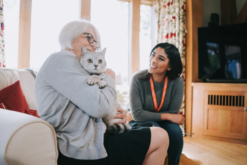 An elderly lady sat on the sofa cuddling her cat, accompanied by her caregiver