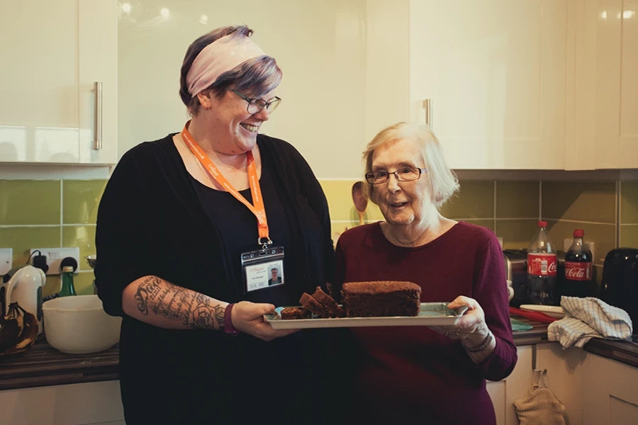 A caregiver with a bright headband holds a tray with a freshly baked cake, standing next to a smiling elderly woman in a kitchen, illustrating the section on what live-in care is in the South West.