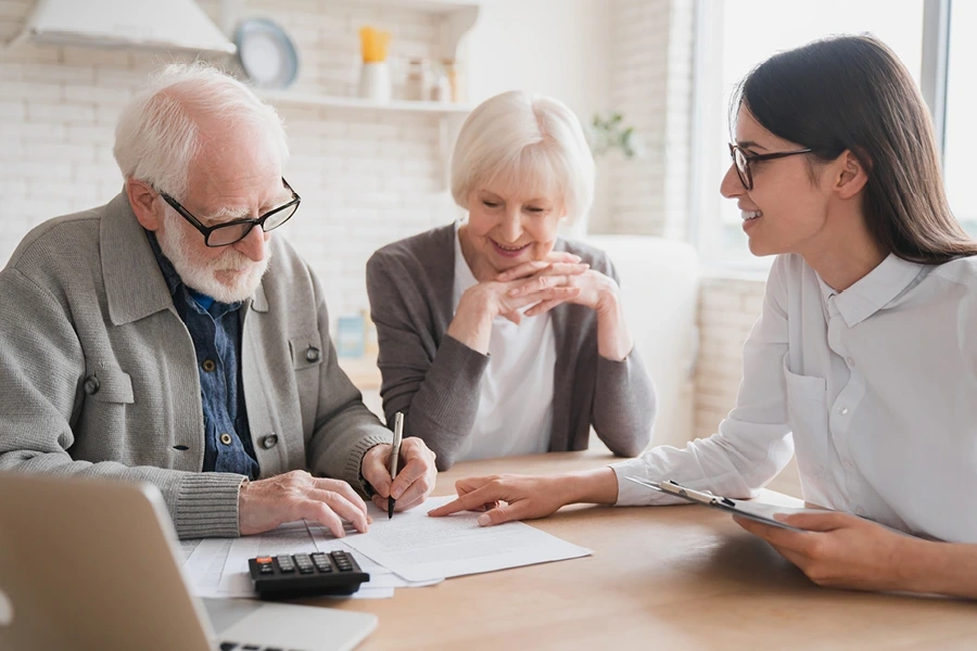 Questions to Ask About Live-in Care in Buckinghamshire, an elderly couple, both with white hair, sit at a table discussing paperwork with a female advisor. The advisor, with long dark hair and glasses, smiles as she points to the documents, while the elderly man signs a paper and the elderly woman looks on with a smile.