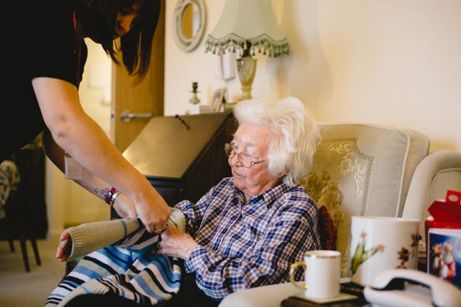 Derbyshire ensuring compatibility in live-in care, showing a caregiver carefully wrapping a cosy striped blanket around an elderly woman seated on a comfortable armchair, highlighting the personalised care and attention to detail in live-in services.