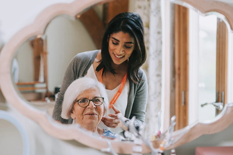 Derbyshire what is live-in care, featuring a caregiver with long dark hair and an orange lanyard, gently assisting an elderly woman with white hair and glasses, smiling as they look into a mirror, showcasing the companionship and support provided in live-in care.