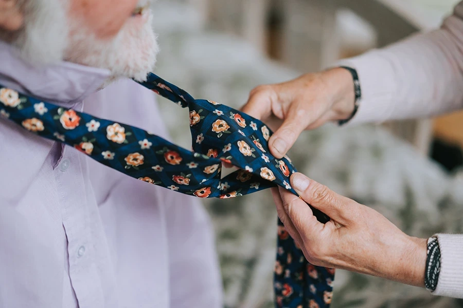 A caregiver helping an elderly man with a patterned tie, illustrating the 'Why Hertfordshire Live-In Costs Vary' section. The elderly man wears a lavender shirt, and the caregiver ensures the tie is correctly adjusted in a close-up shot highlighting the detail.