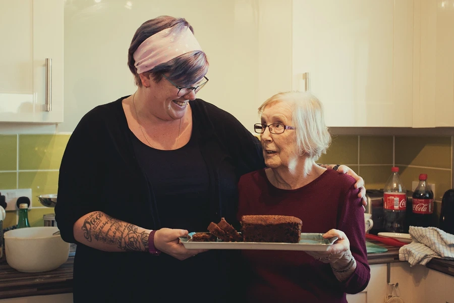 "Wiltshire live-in home care assistant with pink hair and tattoos smiling with elderly client in kitchen, sharing a moment over a freshly baked loaf and slices on a tray.