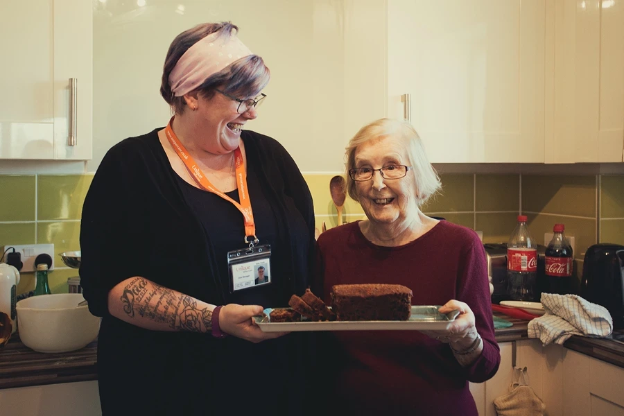 A cheerful caregiver and an elderly woman baking together, depicting the 'Herts Questions to Ask About Live-In Care' section. They hold a tray with a freshly baked cake in a cozy kitchen, with the caregiver wearing a name tag and tattoos visible on her arms.