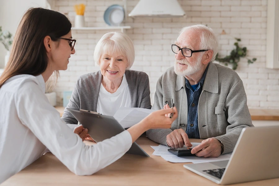 Questions to ask about Derby live-in care, illustrating an elderly couple discussing documents with a young female advisor with long hair, all seated at a wooden table in a bright, modern kitchen, symbolising the need for thorough understanding and clear communication when choosing care options.