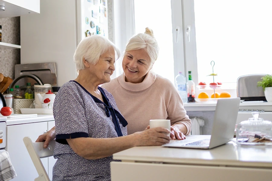 An elderly woman (client) and a middle-aged woman (caregiver) smiling and using a laptop together at a kitchen table, showcasing the section on why live-in costs vary in Warwickshire.