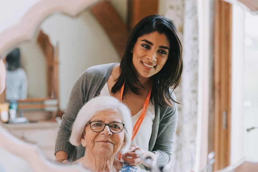 A caregiver helping an elderly woman with her hair, illustrating the 'What is Hertfordshire Live-In Care' section. The caregiver smiles warmly while the elderly woman looks pleased, wearing glasses and a light-colored blouse, with the caregiver wearing a lanyard and badge.