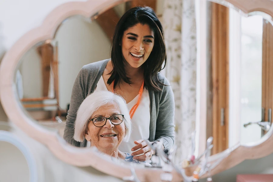 In the 'What is Live-in Care' section for Worcestershire, a cheerful care worker and an elderly woman look at their reflection in a decorative mirror. The elderly woman is wearing glasses and both are smiling, creating a warm and friendly atmosphere.