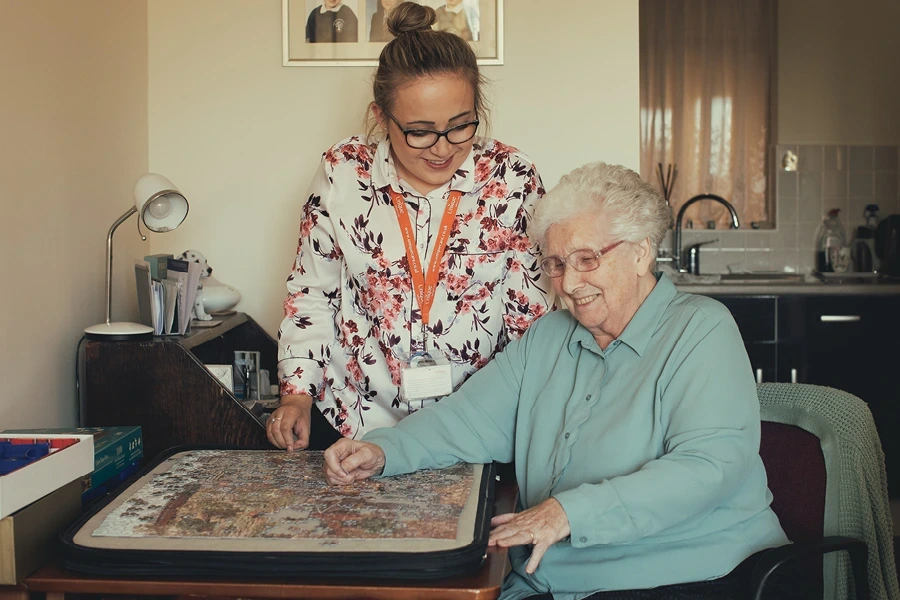 Types of Live-in Care Service in Buckinghamshire, a caregiver with a floral blouse and glasses assists an elderly woman with white hair in completing a jigsaw puzzle. They are seated at a table in a cosy, well-lit room, demonstrating the engaging activities available to clients.