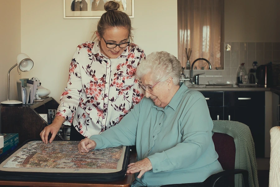 A caregiver with a floral blouse helps an elderly woman with a puzzle in a cosy living room. The background features a kitchenette and family photos. This image pertains to the 'What to Look for in a Live-in Care Provider' section in Oxfordshire.