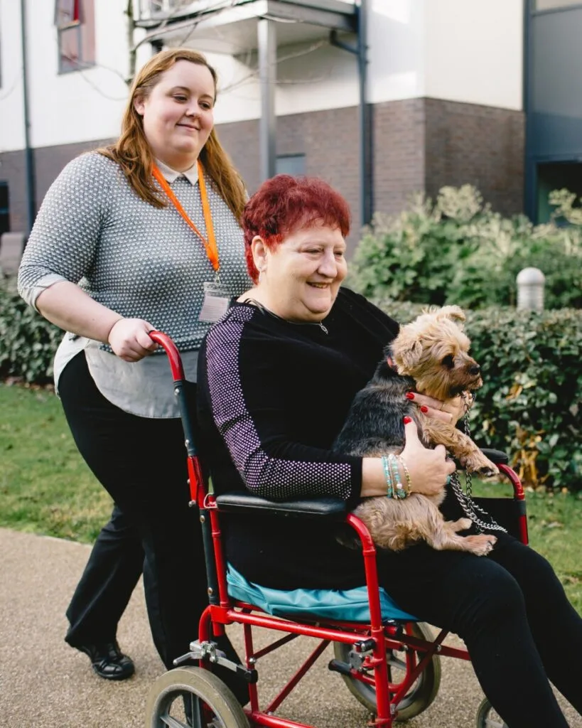 A caregiver is pushing a smiling elderly woman in a red wheelchair outdoors. The elderly woman is holding a small dog on her lap. The background shows a building and greenery.