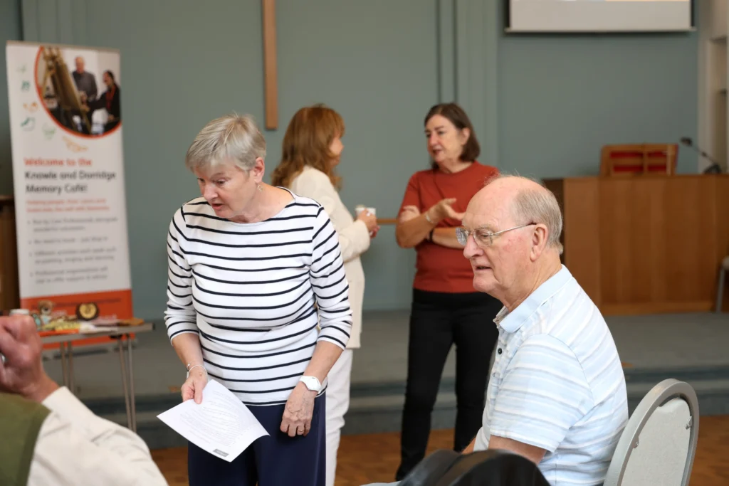 A group of people engaging in conversation at the Family Dementia Workshop, with a woman in a striped shirt speaking to a seated man.