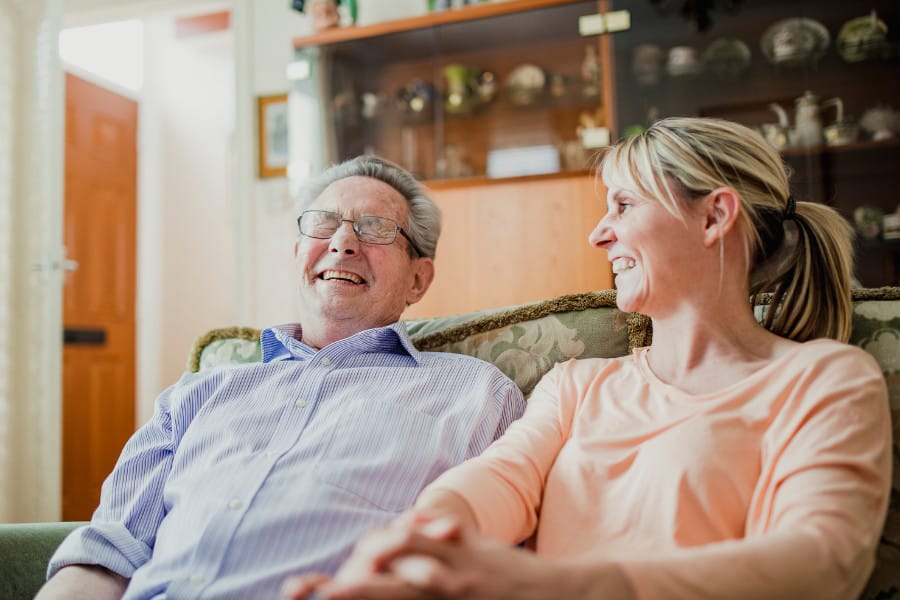 A woman and elderly man sat together on a sofa laughing warmly