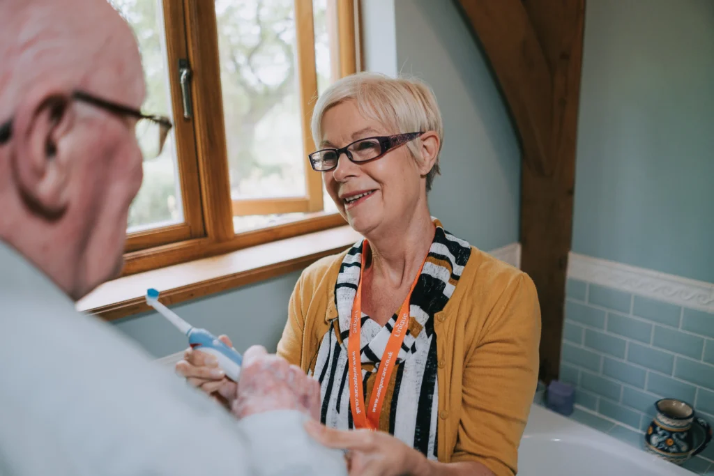 A caregiver smiling while wearing a Unique Senior Care lanyard supporting an older man with brushing his teeth