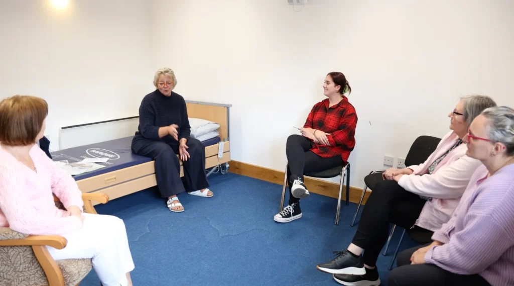 A small group of caregivers participating in a training discussion, seated in a room with a bed for practical demonstrations.