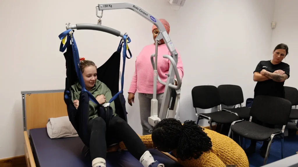 A caregiver demonstrating the use of a hoist to lift and support a trainee in a practical training session