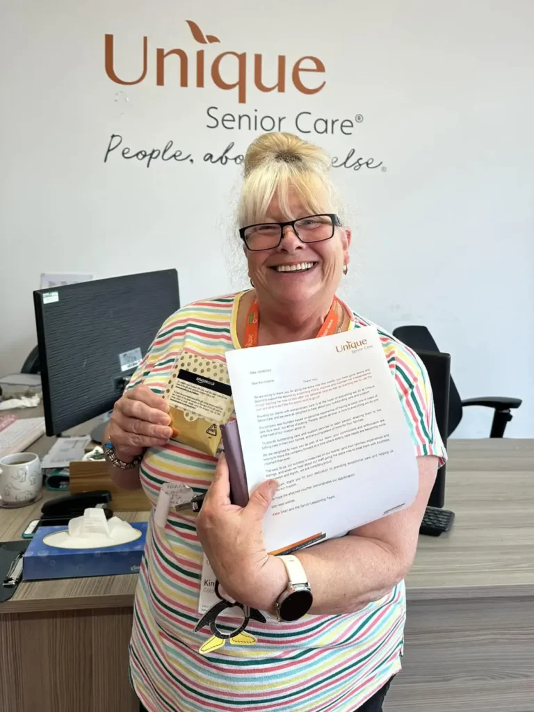 A smiling caregiver holding her recogntion award and a gift while standing in front of the Unique Senior Care logo at an office desk.