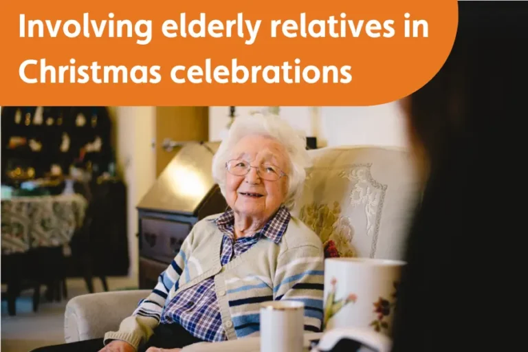 “An elderly woman with glasses smiles warmly while sitting on a sofa, enjoying a cosy conversation. The room has a relaxed, festive atmosphere, and a mug of tea sits nearby.