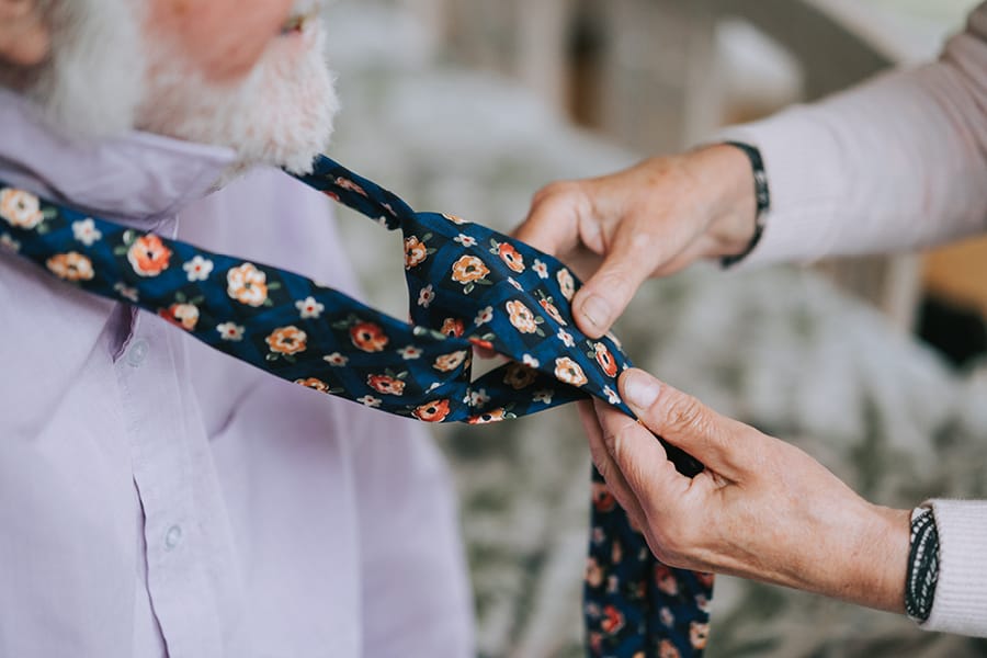 Female unique caregiver assisting older gentleman client with his neck tie in the types of yorkshire live in care page.