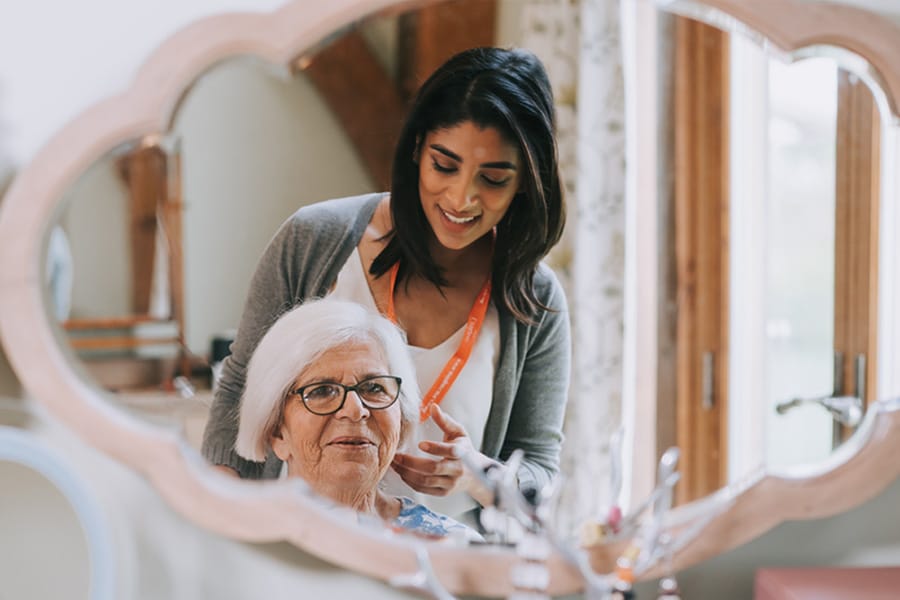 Elderly lady with white hair with her Caregiver stood behind her both looking into and ornate shaped mirror in the what is live in care section of the Yorkshire page.