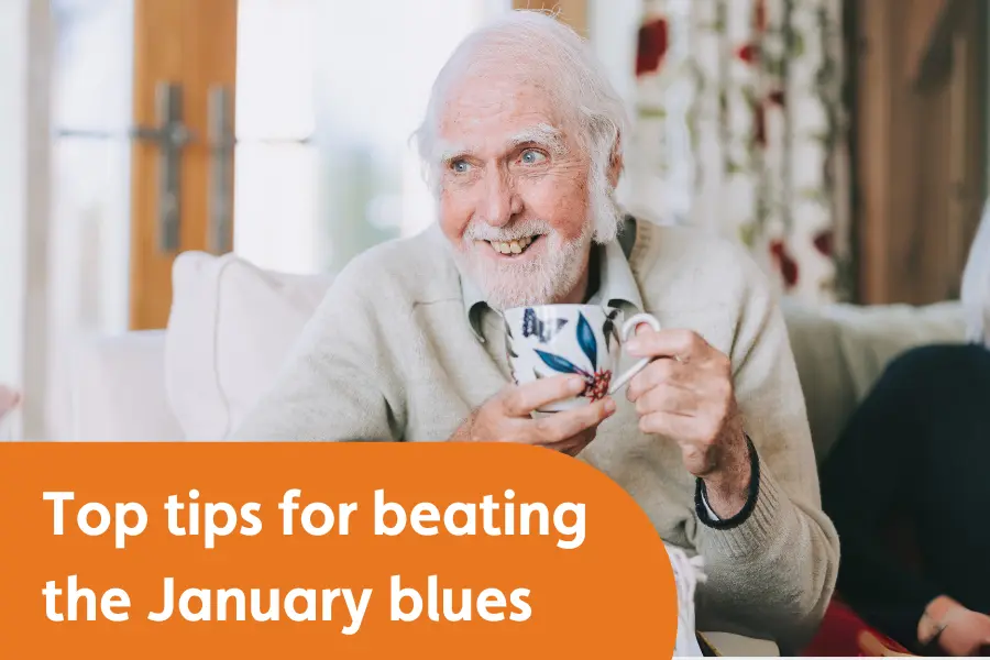 An elderly man with white hair and a beard smiles warmly while holding a patterned mug. He is sitting comfortably on a sofa in a cosy, well-lit living room. Below him, an orange banner reads, "Top tips for beating the January blues."