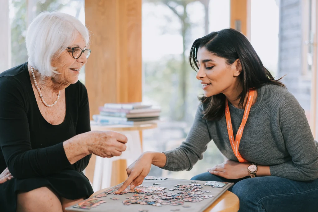 Elderly woman and caregiver with an orange lanyard solving a jigsaw puzzle together in a bright room.